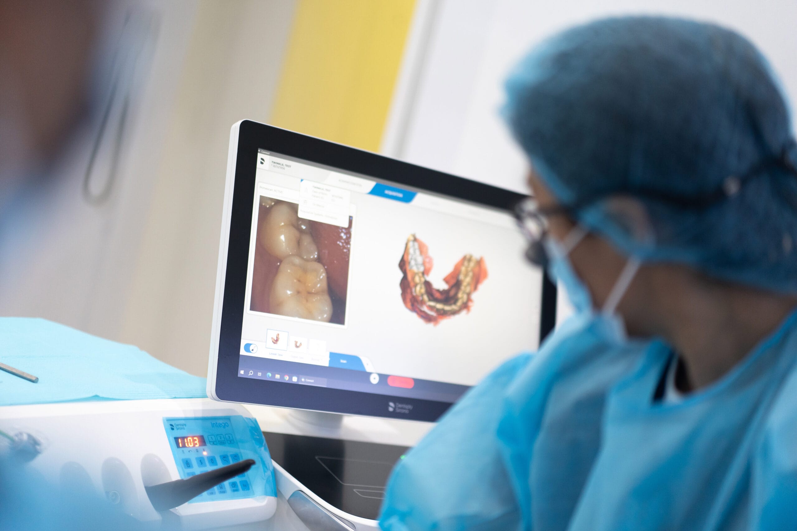 A dental professional in blue surgical gear reviewing digital intraoral scans and photos of a patient's teeth on a computer monitor.