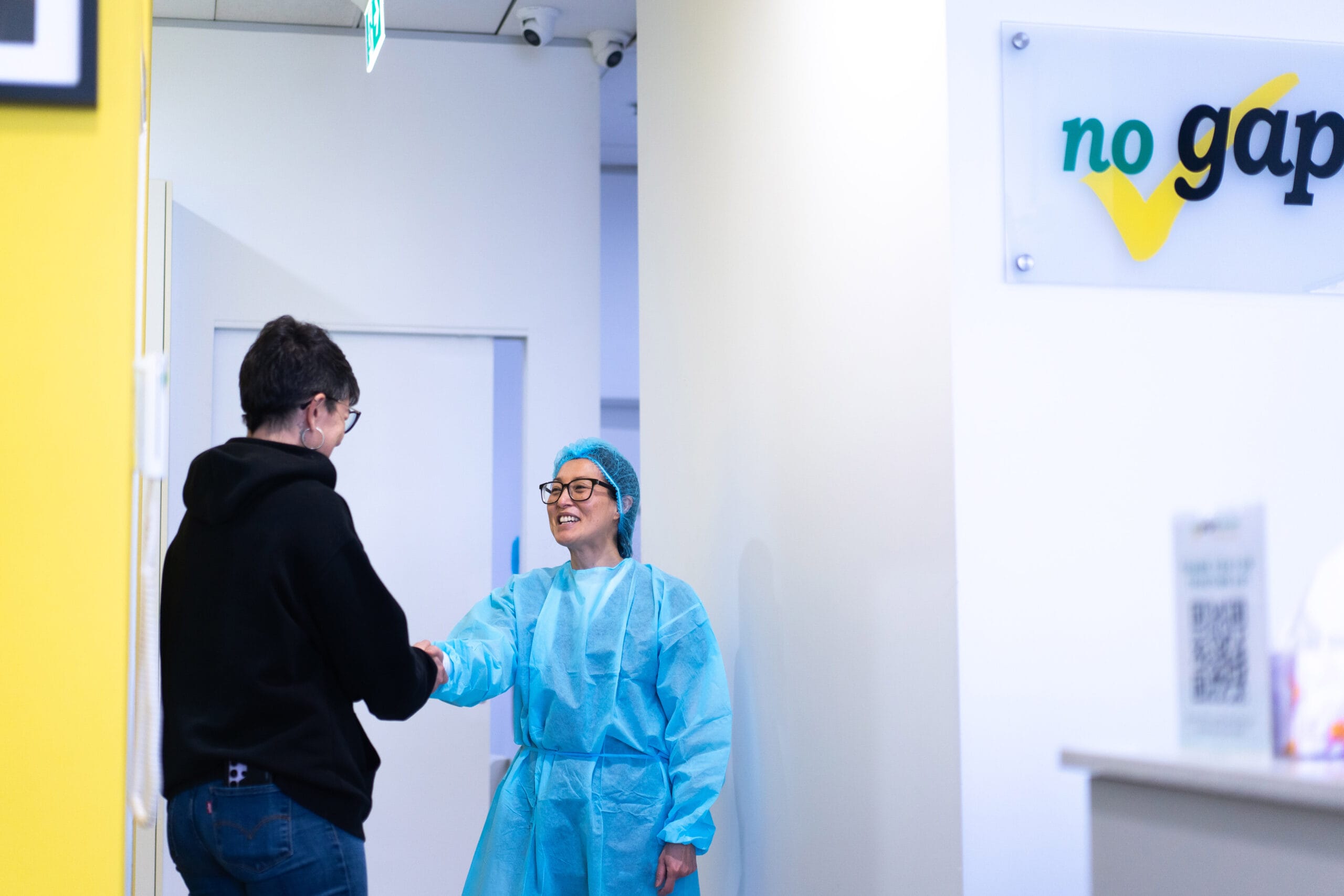 A female dental professional in blue surgical gear and a hairnet smiling while shaking hands with a patient at a No Gaps Dental clinic.