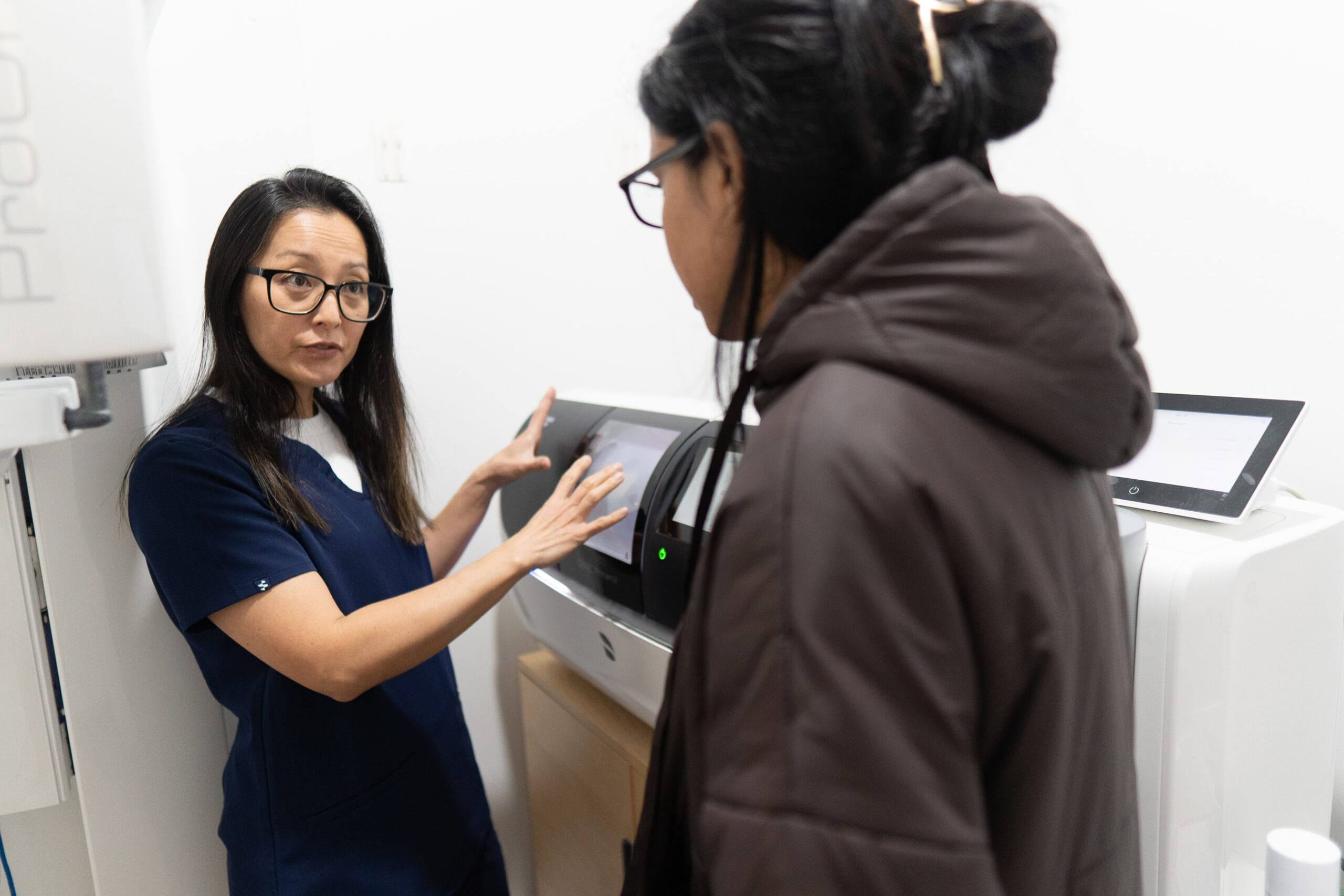 A dental professional in dark blue scrubs explaining a piece of digital medical equipment to a patient in a brown jacket.