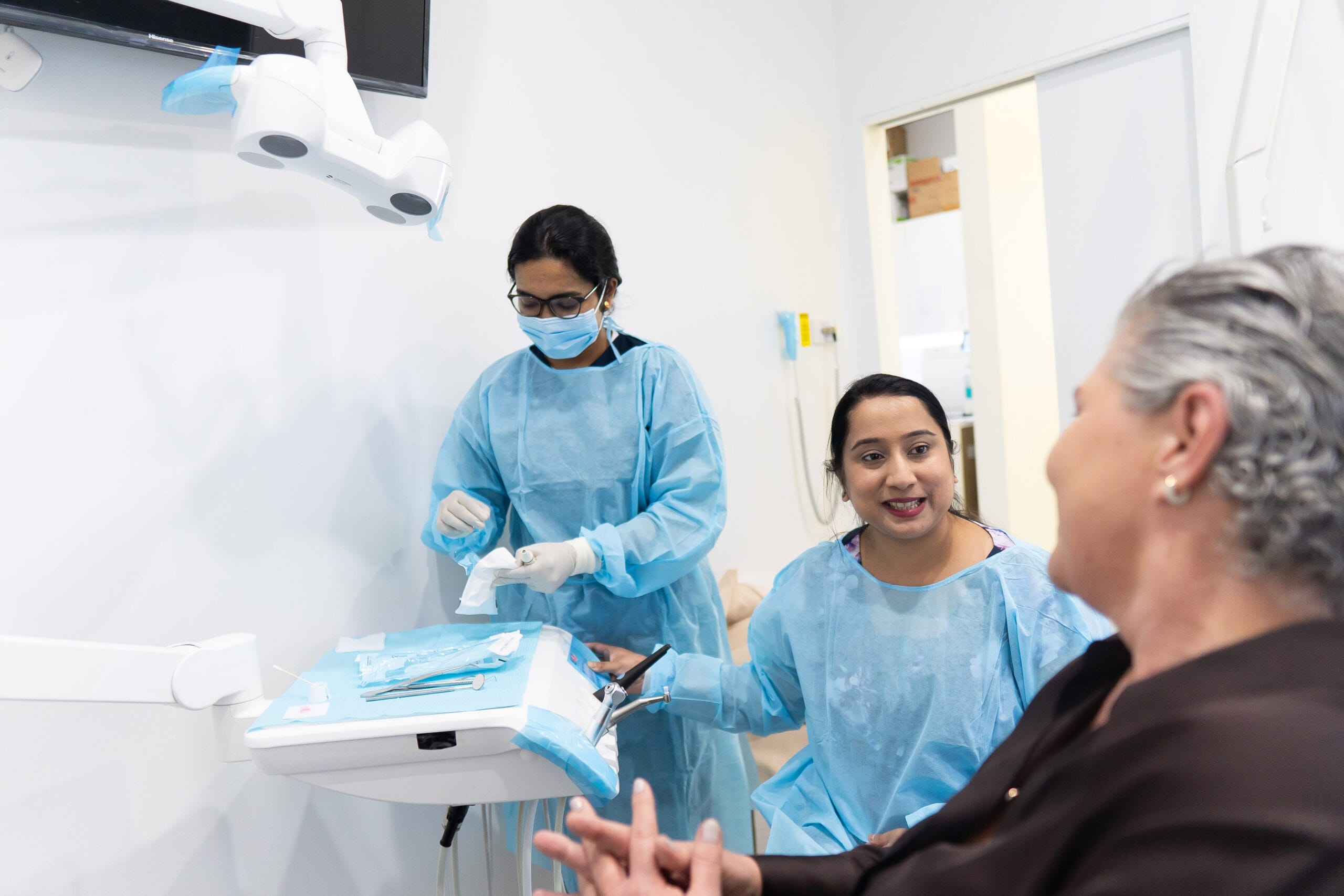 wo dental professionals in blue scrubs and face masks talking with a seated patient in a bright clinical treatment room.