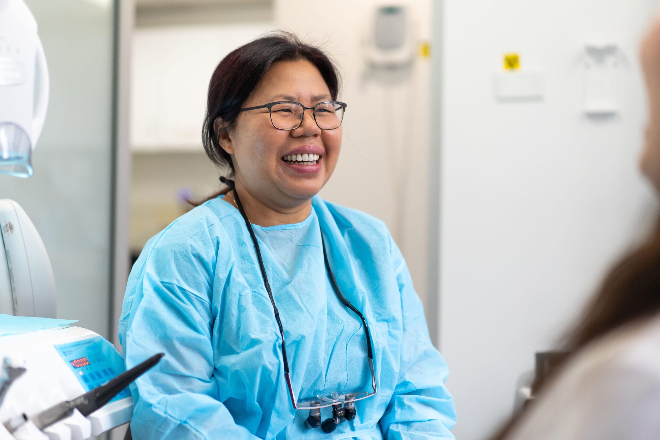 A smiling female dentist in blue scrubs and glasses laughing during a patient consultation in a treatment room.