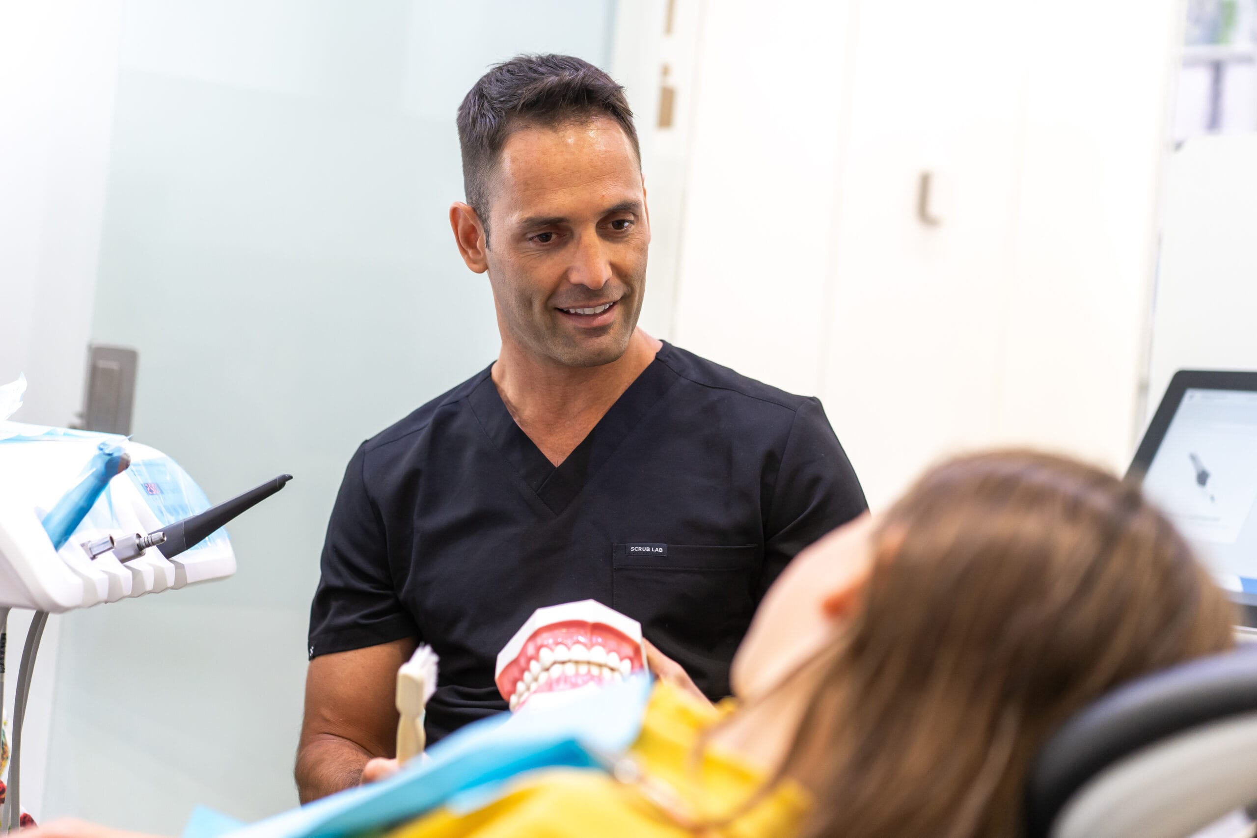 A dentist in a black scrub top explaining a procedure to a patient using a dental model.