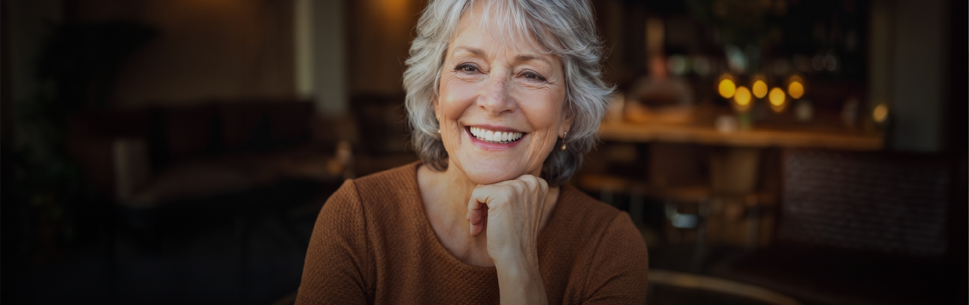 Smiling older woman with gray hair in a brown sweater sits in a cozy cafe, chin resting on her hand.