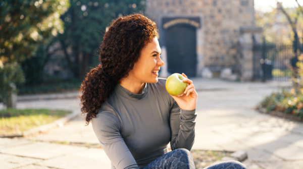 Woman eating green apple | No Gaps Dental Smiling woman sits outdoors in a park, holding a green apple near her face as she enjoys a sunny day.