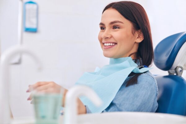 Woman in dental chair | No Gaps Dental Woman sitting in a dentist's chair, smiling with a light blue dental bib on, hands holding a cup (patient during checkup).